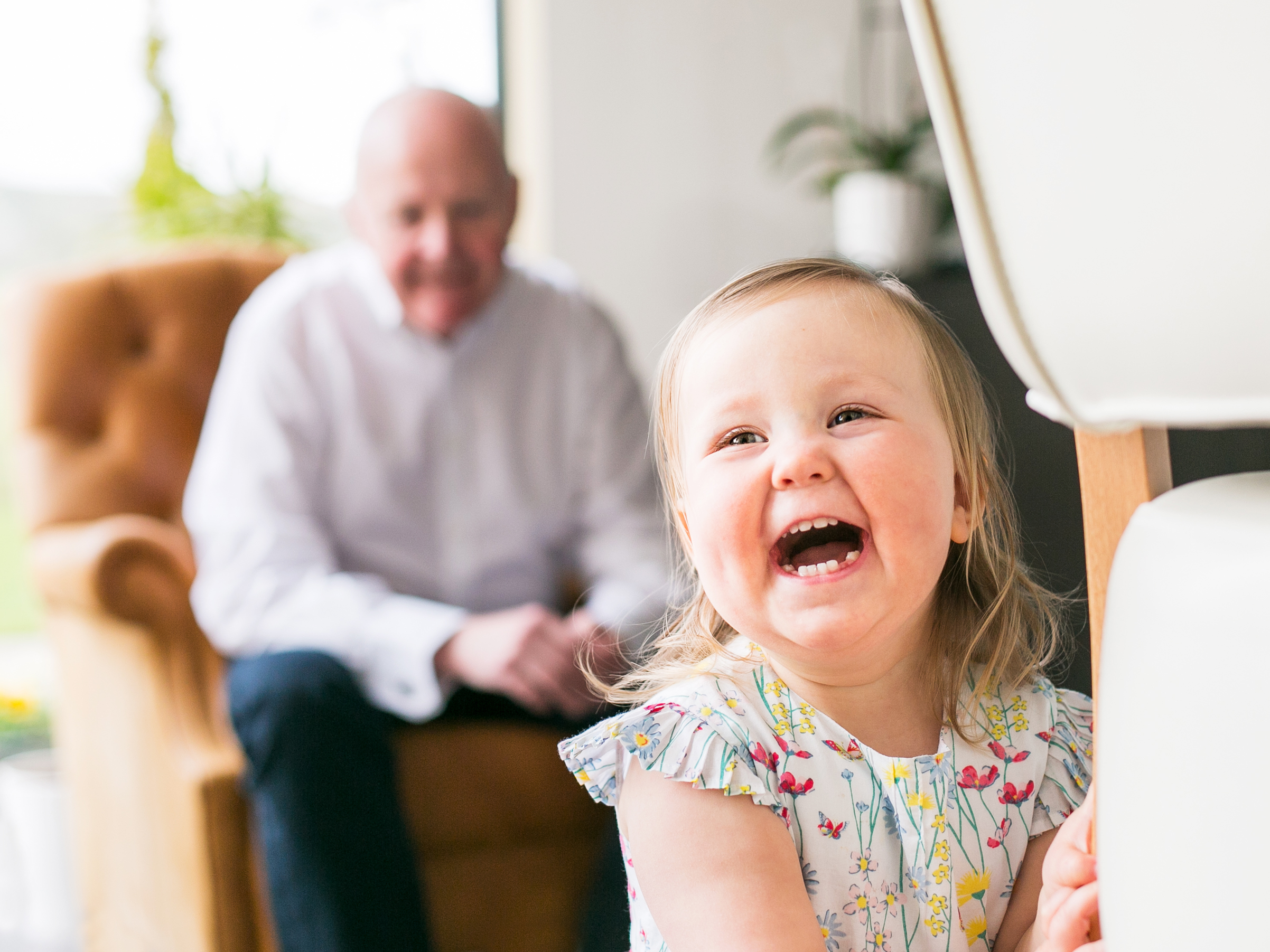 Colour photo of a little girl laughing at the camera with grandad in the background