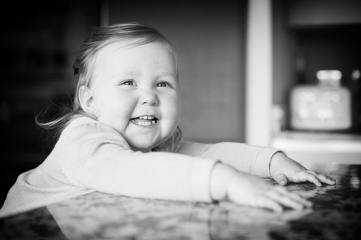 B&W photograph of smiling 2 year old girl sat in a kitchen