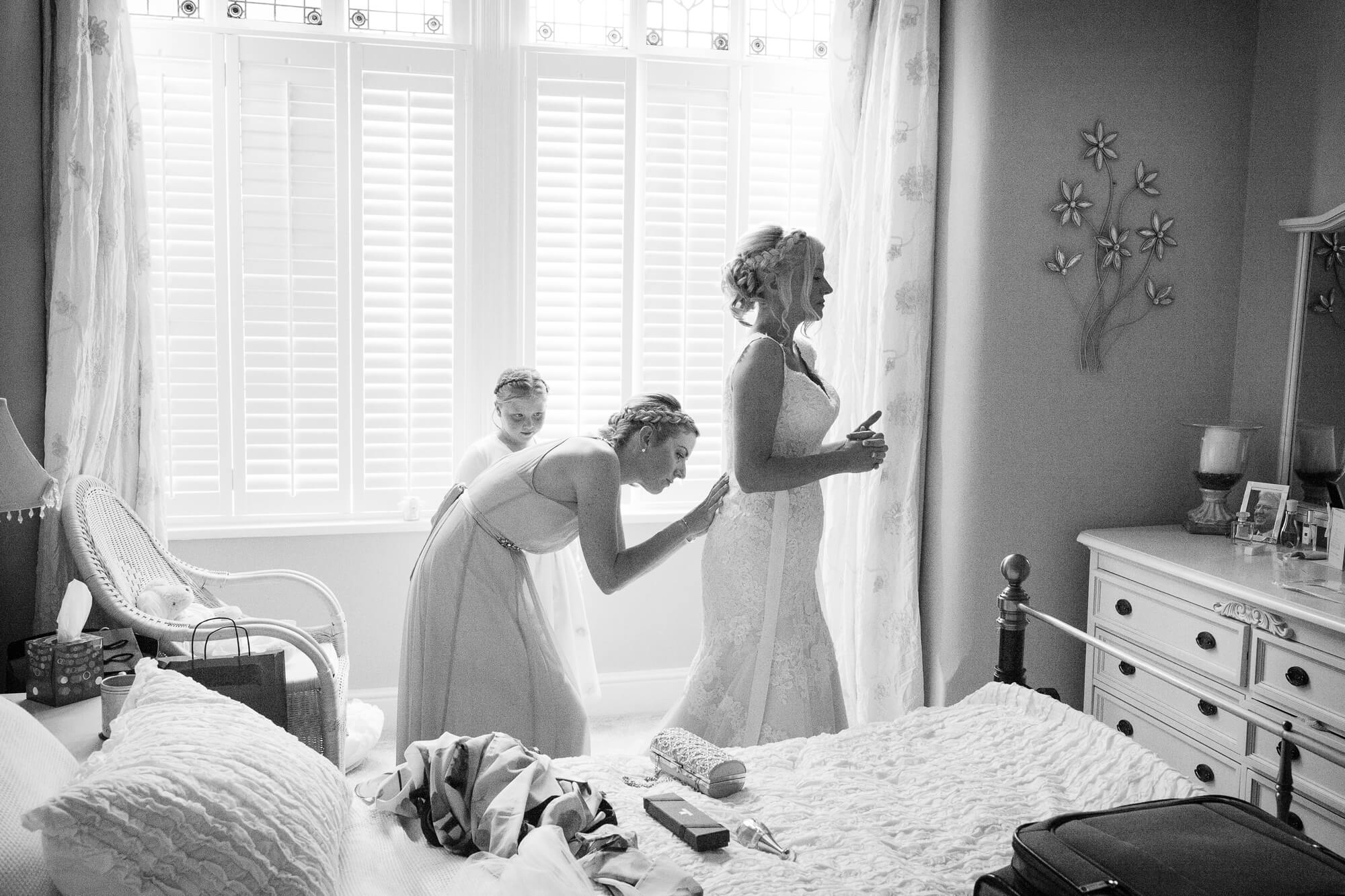 Black & white moody image of bridesmaid fastening brides dress in bedroom