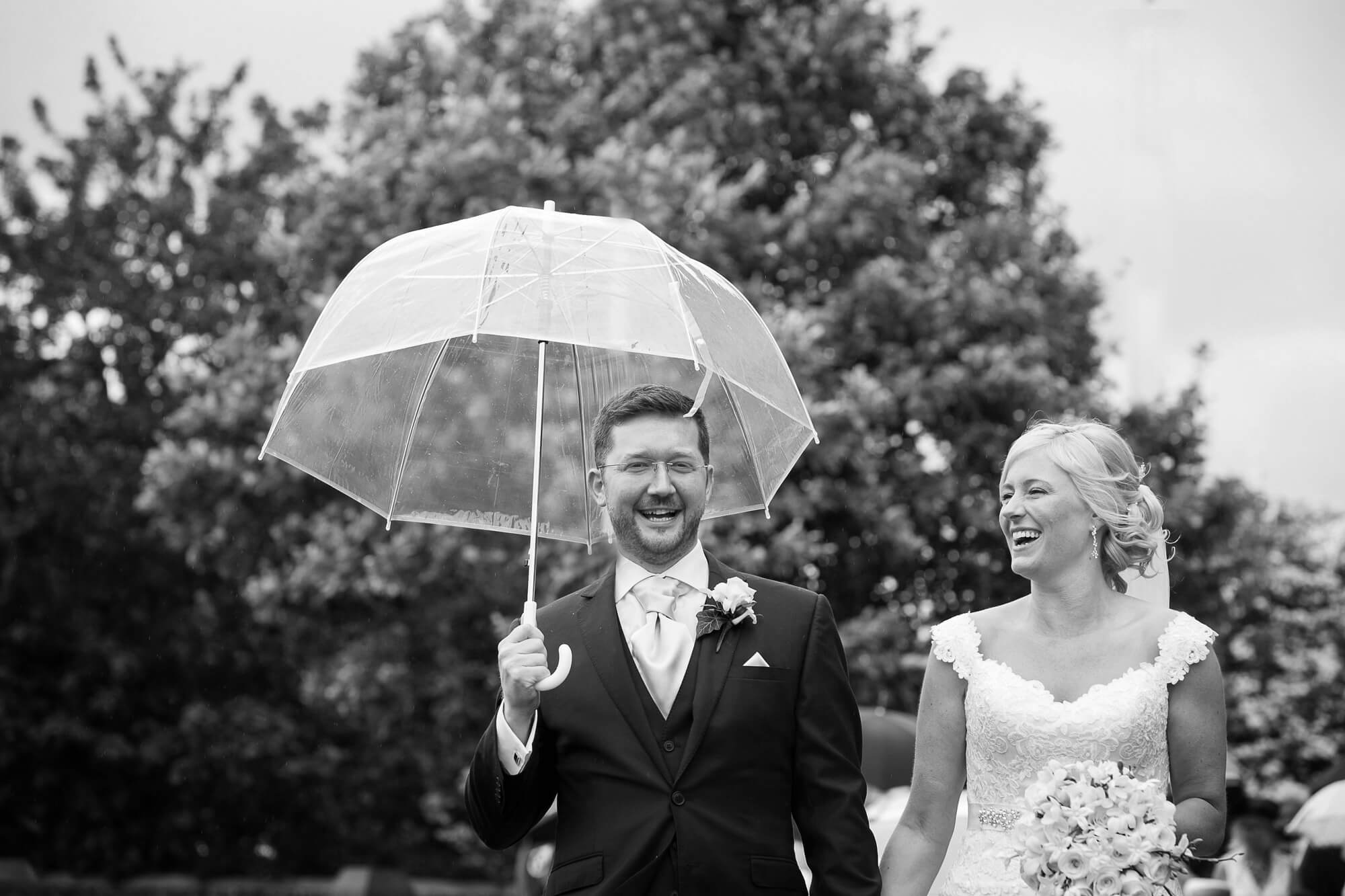 Black & white photo groom holding clear umbrella, bride laughing as they leave church