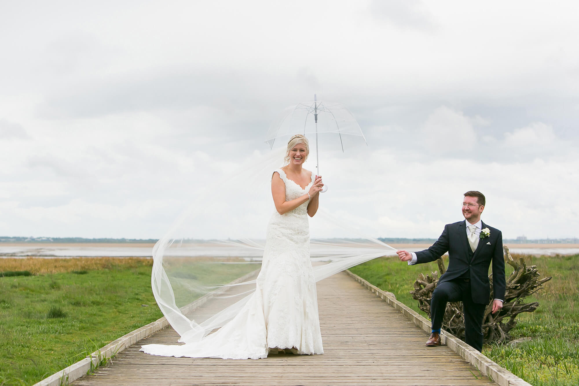 colour photo of bride & groom with umbrella in the wind. Dress blowing. On beach