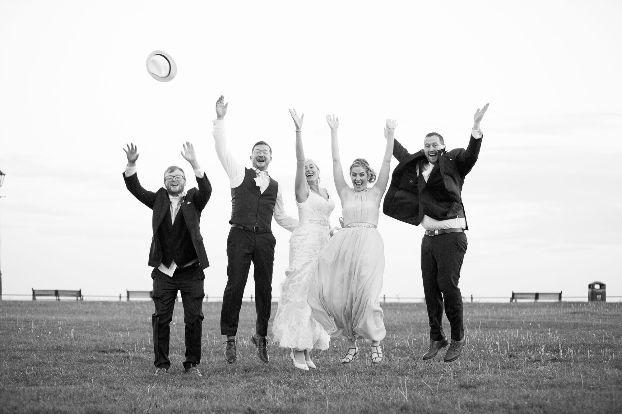 black & white image of bride, groom, best man nad bridesmaids throwing hats in the air and jumping on the promenade
